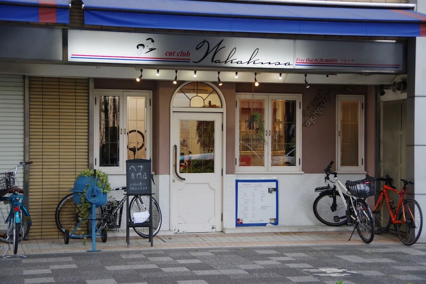 La tienda tiene un letrero que dice "cut club Wakana" en la parte superior de una puerta doble blanca, y ofrece servicios de atajos y henna. Intercalado entre las ventanas y el estacionamiento de bicicletas, hay portabicicletas azules, plantas de interior y menús y letreros cerca de la entrada.