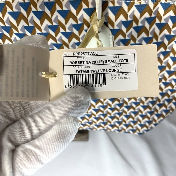La mano enguantada sostiene la etiqueta del producto "ROBERTINA 2 (DUE) SMALL TOTE" en la parte superior de la tela con estampado de tatami con un patrón geométrico en azul, marrón y blanco, que es el estilo característico de la colección de bolsos pequeños de ROBERTA PIERI.