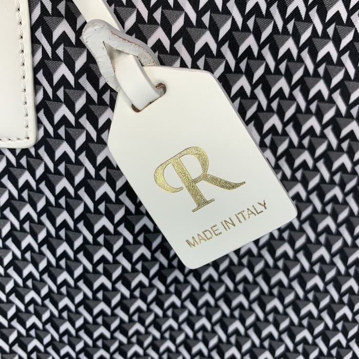 Una etiqueta ROBERTA PIERI de cuero blanco con las letras "R" en oro y las palabras "MADE IN ITALY" cuelga del fondo de una tela geométrica en blanco y negro.