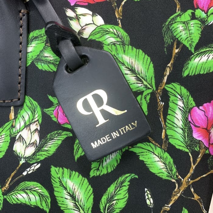 La etiqueta de equipaje negra ROBERTA PIERI con la "R" dorada y el texto italiano está suspendida de una tela floral con hojas verdes y flores rosas, complementando la mini bolsa de lona con la elegancia italiana atemporal.