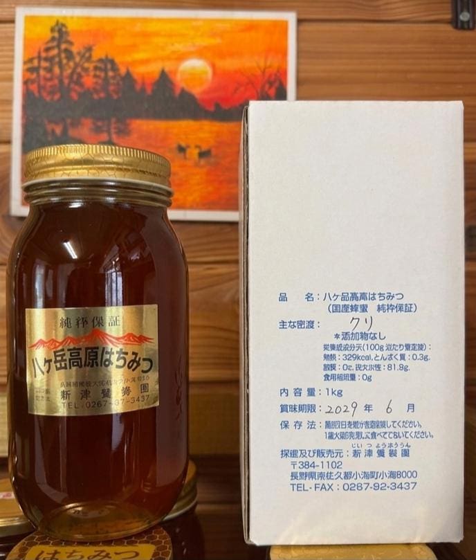 「八ヶ岳高原 国産天然はちみつ 1kg」の瓶には「純粋保証」のラベルが貼られ、ハチミツの種類として「クリ（日本の栗）」と記されており、青い日本語の文字が施された白い箱の横に置かれている。 その背後には、八ヶ岳高原の静謐な風景を彷彿とさせる、木々や山々を描いたオレンジ色の夕焼けの絵が、木製の台の上に絵のように美しい背景を演出している。.
